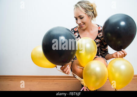 Beau modèle assis sur un plancher en bois en se penchant en arrière contre le mur blanc d'oeil tête hors de ballons d'or et noir Banque D'Images