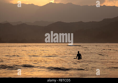 Soleil derrière les montagnes de Na Pali avec une silhouette de surfer dans l'avant-plan et le soleil reflétant sur l'océan Banque D'Images