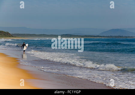 Un jeune surfeur solitaire se promène le long du bord de l'eau avec sa planche de surf sur une plage de la côte sud de Nouvelle-Galles du Sud Banque D'Images