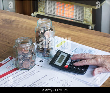 Dame âgée dans ses années 90 regardant des factures de gaz, électriques, de services publics avec calculatrice et des pots de pièces sur table. Incendie de gaz en arrière-plan Banque D'Images