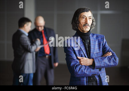 Ancien businessman wearing blue Jacket debout avec les mains jointes. L'arrière-plan à deux hommes en costumes discuter de documents. Portrait de la haute direction, la retraite formalités. Immeuble de bureaux à l'intérieur. Banque D'Images