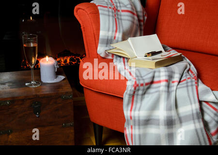 Douce couverture, livre, le bloc-notes et stylo sur un élégant fauteuil, un verre de vin et la bougie à côté et cheminée en arrière-plan Banque D'Images