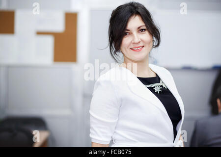 Succès attractive brunette avec genre yeux debout dans la salle de conférence et sourire accueillant. Vêtus de blanc, veste robe noire stricte des bijoux. Portrait of cute young business woman posing. Banque D'Images