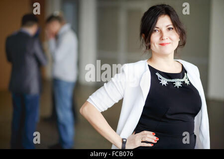 Brunette avec succès patron attrayant genre yeux se tient à l'intérieur de bâtiment de bureaux et sourire accueillant. Une stricte robe noire bijoux. Jeune Chef d'entreprise woman posing. En arrière-plan l'homme de discuter. Banque D'Images
