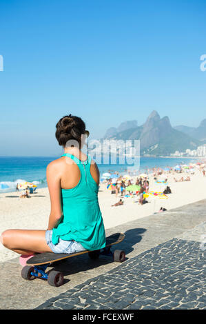 Jeune femme brésilienne assise sur son skateboard donnant sur la plage d'Ipanema à Arpoador à Rio de Janeiro, Brésil Banque D'Images