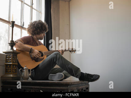 Loft vivant. Un jeune homme qui joue de la guitare assis par une fenêtre. Banque D'Images