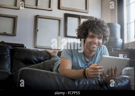 Décoration Loft. Un mur décoré de photos dans des cadres, à l'envers pour montrer le dos. Un homme allongé sur un canapé, à l'aide d'une tablette numérique. Banque D'Images