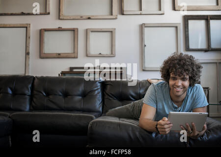 Décoration Loft. Un mur décoré de photos dans des cadres, à l'envers pour montrer le dos. Un homme allongé sur un canapé, à l'aide d'une tablette numérique. Banque D'Images