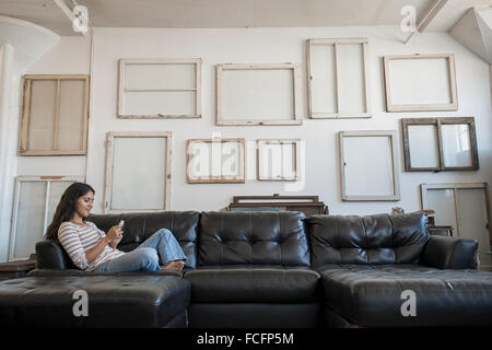 Décoration Loft. Un mur décoré de photos dans des cadres, à l'envers pour montrer le dos. Une femme assise sur un canapé à l'aide d'un téléphone intelligent. Banque D'Images