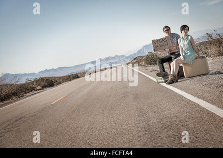 Un jeune couple, homme et femme, sur une route goudronnée dans le désert hitchiking, avec un panneau disant Vegas ou buste. Banque D'Images
