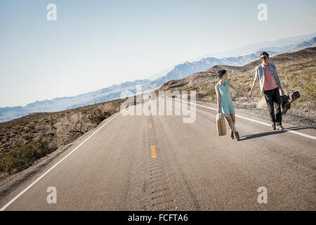 Un jeune couple, homme et femme, marche main dans la main sur une route goudronnée dans le désert des étuis de transport. Banque D'Images