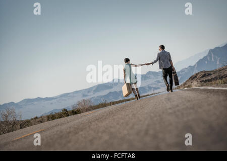 Un jeune couple, homme et femme, marche main dans la main sur une route goudronnée dans le désert des étuis de transport. Banque D'Images