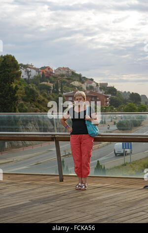 Hauts smiling blonde woman in red Pantalon et chemisier noir est debout sur la passerelle au-dessus de la route à Santa Susanna, Catalogne, Banque D'Images