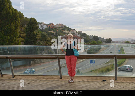 Hauts smiling blonde woman in red Pantalon et chemisier noir est debout sur la passerelle au-dessus de la route à Santa Susanna, Catalogne, Banque D'Images