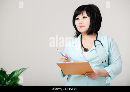 Infirmière en courbes, est encore aujourd'hui par écrit les résultats stylo à papier, en blouse de laboratoire médical avec stéthoscope autour de son cou. Apparence asiatique, cheveux courts. La végétation petite sur le premier plan. Banque D'Images