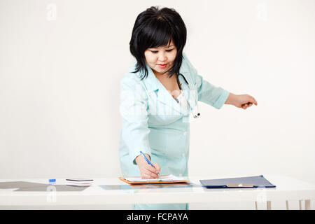 Curvy mignon femme médecin à son bureau permanent écrit Résultats du sondage sur le papier par la plume, manteau de laboratoire médical avec stéthoscope autour du cou. Apparence asiatique, cheveux courts. Banque D'Images