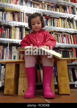 Jolie petite fille avec un manteau et bottes en train de lire un livre épais à bookshop Banque D'Images