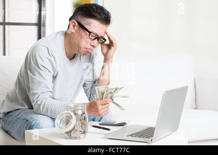 Problème financier concept. Portrait de 50s mature Asian man comptant de l'argent avec l'expression inquiète, assis sur le canapé à la maison. Banque D'Images