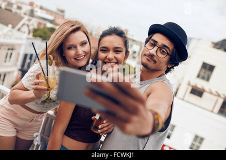 Les personnes bénéficiant de multiraciale en partie avec des boissons et prendre un autoportrait. Trois jeunes amis en tenant avec selfies smart phone Banque D'Images