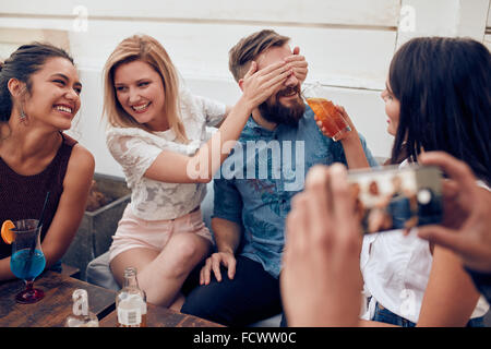 Les jeunes gens assis jouissant ensemble partie. Femme fermeture yeux d'un homme avec un autre donner à boire. Jeunes amis de s'amuser à Banque D'Images