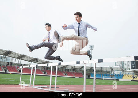 Deux hommes d'affaires de courir et de sauter sur des obstacles sur une piste avec l'arrière-plan de terrain de sport Banque D'Images