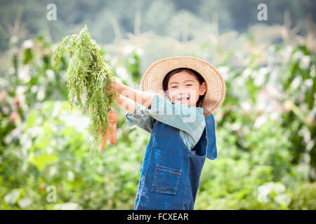 Fille dans un chapeau de paille holding carrots up and smiling at field Banque D'Images