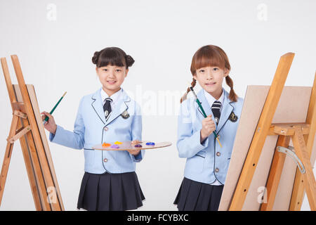 L'école élémentaire deux fille dans la classe d'art tenant un pinceau et une palette avec un chevalet chaque fois regardant en avant Banque D'Images