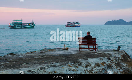 Homme seul est assis sur un banc sur la côte en regardant les bateaux de pêche au coucher du soleil. Banque D'Images