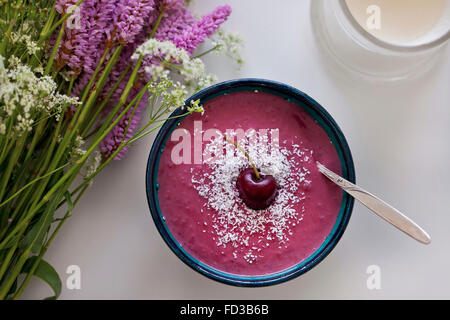 Cerisier rose et noix de coco smoothie dans un bol, surmontée de la noix de coco râpée et de cerise fraîche, violet et blanc fleurs sur la table Banque D'Images