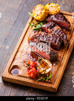 Tranches de steak Ribeye avec le maïs et tomates cerises sur une planche à découper sur fond de bois Banque D'Images