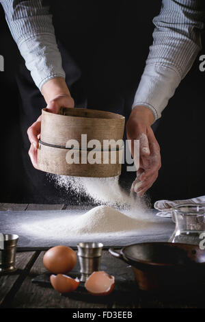 Les mains de farine tamisage tamis ancien sur ancienne en bois table de cuisine. Vintage ustensiles avec de la farine, de l'eau et les œufs au foregro Banque D'Images