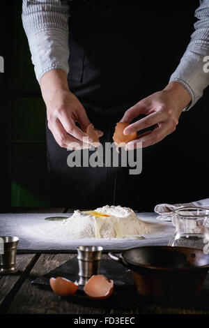 Femmes casser l'œuf en farine pour les pâtes sur la vieille table de cuisine en bois. Ustensiles Vintage et oeufs cassés au premier plan. Banque D'Images