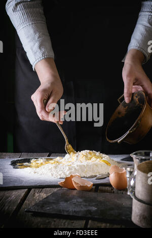Les mains dans la farine les oeufs pétrir des pâtes par fourchette sur ancienne en bois table de cuisine. Oeufs cassés au premier plan. Style rustique foncé. Banque D'Images