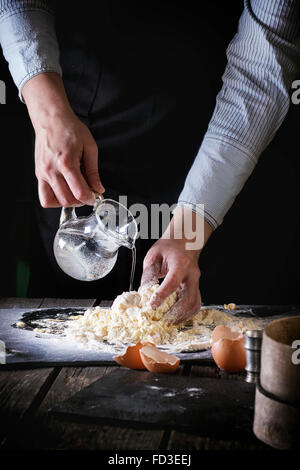 Les mains des femmes de l'eau courante à partir de la verseuse, pétrir la pâte pour les pâtes sur la vieille table de cuisine en bois. Oeufs cassés au premier plan. La rouille noire Banque D'Images