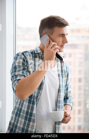 Profil de l'homme séduisant de boire du café et conversation sur téléphone cellulaire près de la fenêtre Banque D'Images