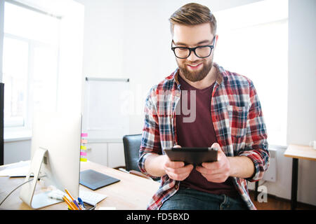 Happy handsome man with beard en chemise à carreaux et des lunettes assis sur la table dans le bureau et à l'aide de tablet Banque D'Images