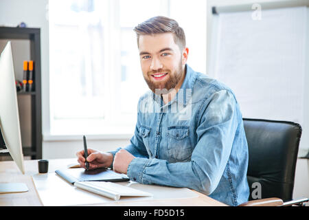 Portrait of happy young designer en jeans shirt travaillant à l'aide de tablette avec stylet in office Banque D'Images