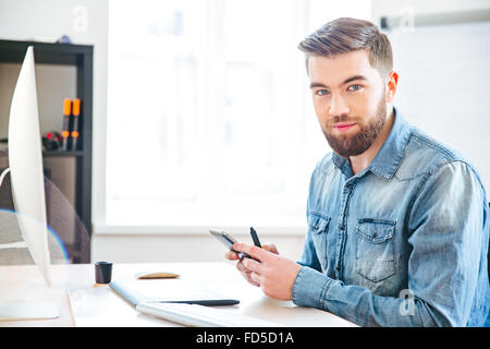 Beau jeune homme barbu souriant assis sur au travail et à l'aide de mobile phone Banque D'Images