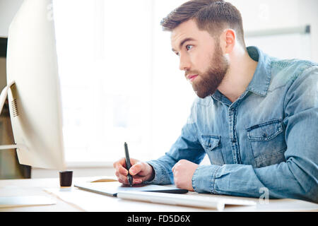 Beau jeune designer concentré avec barbe en bleu shirt dessin à l'ordinateur et tablette graphique au bureau Banque D'Images