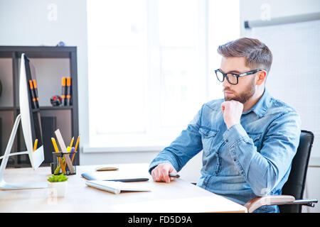 Thoughtful beau jeune designer avec barbe, dans les verres à l'aide de tablette graphique et l'ordinateur et la pensée Banque D'Images