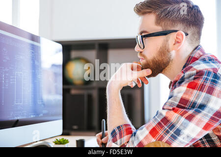 Profil de l'homme barbu concentré dans les verres assis et le dessin des plans sur ordinateur Banque D'Images