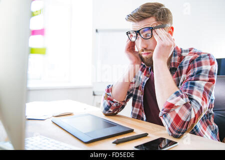 Fatigué épuisé jeune homme avec barbe dans les verres assis sur lieu de travail et de toucher des temples Banque D'Images