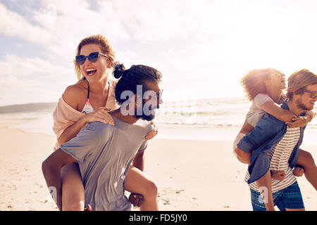 Heureux les jeunes hommes donnant piggyback ride à la femme sur la plage. Groupe varié de jeunes s'amusant sur la plage et de ferroutage. Banque D'Images