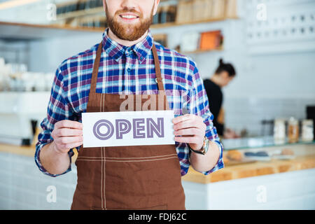 Smiling waiter barbu en tablier brun montrant open sign au coffee shop Banque D'Images