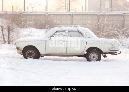 Voiture abandonnée couverte de neige en hiver au coucher du soleil, des tons chaleureux, vue de côté. La rouille, le recyclage, le traitement des métaux, la radiation de l'automobile. Banque D'Images