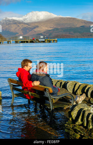 Le Loch Lomond, au Royaume-Uni. 3 Février, 2016. Après les récentes tempêtes et fortes pluies pour la saison, le niveau d'eau à Luss sur le Loch Lomond a surmonté les murs provoquant des inondations locales.Sur la première journée ensoleillée, Tom Wilson et la Géorgie à partir de Londres, en vacances près de Luss, prendre une promenade relaxante pour admirer la neige surmonté Ben Lomond et les eaux calmes du Loch Lomond. Credit : Findlay/Alamy Live News Banque D'Images