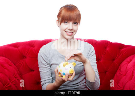 Belle jeune fille aux cheveux rouge mis coin in piggybank sur canapé rouge in front of white background Banque D'Images