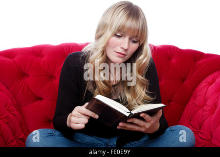 Jeune fille aux cheveux blonds sur canapé rouge lire un livre in front of white background Banque D'Images