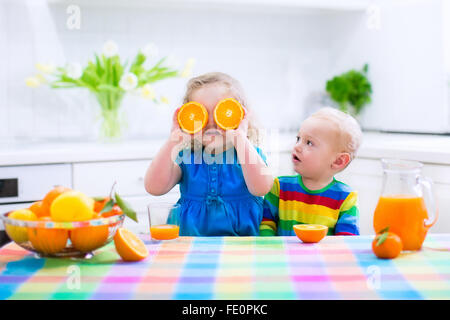Drôle mignon et adorable petite fille garçon bébé boire du jus d'orange fraîchement pressé pour petit-déjeuner sain dans une cuisine blanche Banque D'Images