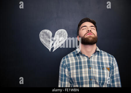 Portrait de jeune homme désespérément malheureux debout avec les yeux fermé plus de tableau noir avec fond tirées cœur brisé Banque D'Images